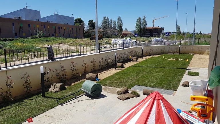 Construction site with green lawn, concrete foundation, metal pipes, and red and white striped barriers under clear blue sky
