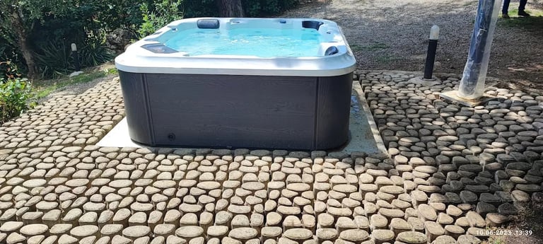 Outdoor hot tub with light blue water and dark grey sides on a paved stone patio next to landscaping